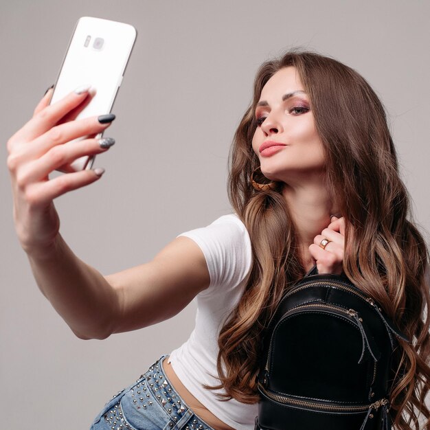 Widok z przodu całkiem młoda dziewczyna z modnym plecakiem robi selfie na telefonie Atrakcyjny model w białej koszuli i dżinsach pozowanie na szarym tle na białym tle w studio Koncepcja casualowego stylu życia