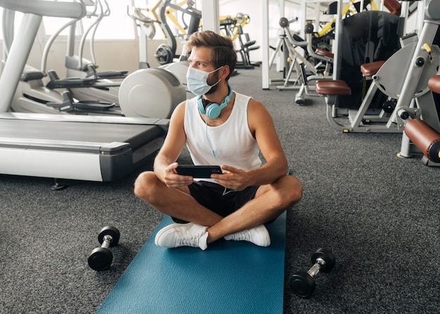 Widok z przodu człowieka ze słuchawkami i maską medyczną, trzymając smartfon na siłowni