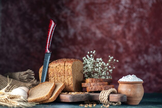 Widok z przodu dietetycznego czarnego chleba pszenicy na drewnianej desce do krojenia nóż kwiat jajka mąka w misce brązowy ręcznik na tle mieszanych kolorów