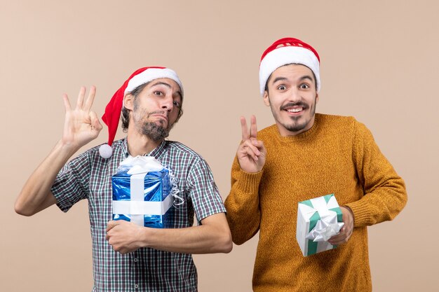 Widok z przodu dwóch błogich facetów, jeden robiący znak okey, a drugi znak zwycięstwa i trzyma prezenty świąteczne na beżowym tle na białym tle