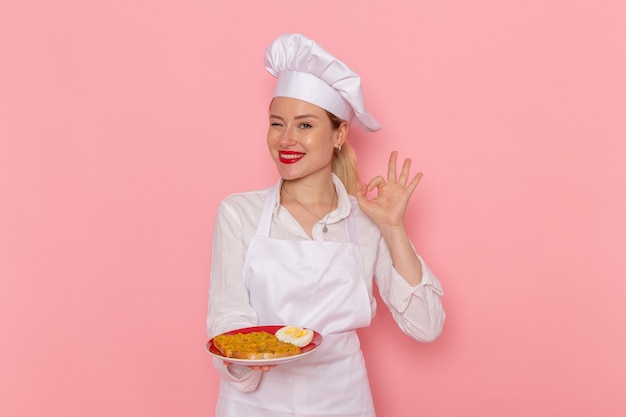 Widok z przodu kobieta cukiernik w białym zużyciu trzymając talerz z jedzeniem na różowym biurku kucharz praca kuchnia kuchnia jedzenie