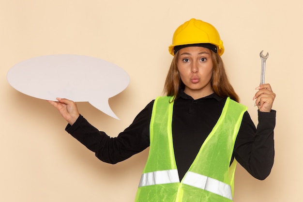 Widok z przodu kobieta konstruktor w żółtym kasku trzymająca duży biały znak ze srebrnym narzędziem na białej ścianie kobieta