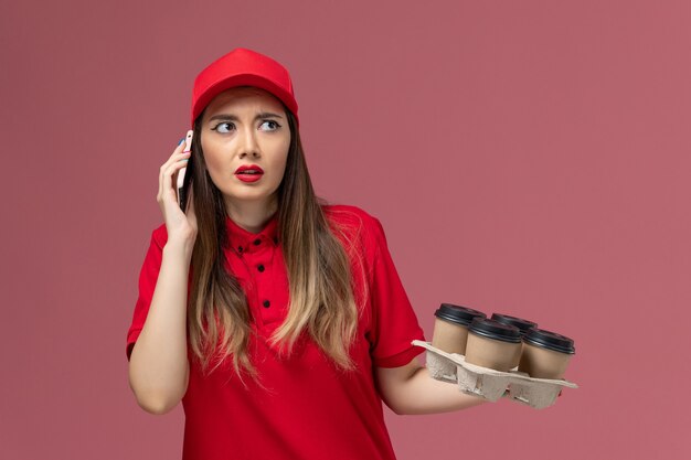 Widok z przodu kobieta kurier w czerwonym mundurze, trzymając filiżanki kawy dostawy rozmawia przez telefon na różowym tle mundur dostawy usług