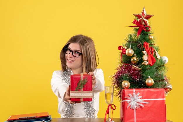 Widok z przodu kobieta lekarz siedzi przed stołem z prezentami i drzewem na żółtym tle z choinką i pudełkami na prezenty