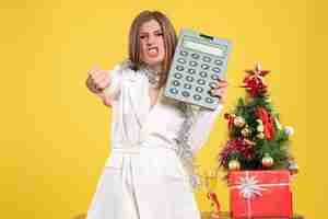 Bezpłatne zdjęcie widok z przodu kobieta lekarz stojący i trzymając kalkulator na żółtym tle z choinką i pudełkami na prezenty
