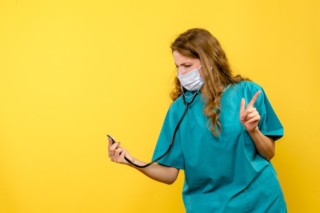 Widok z przodu kobiety lekarz w masce za pomocą stetoskopu na żółtej ścianie