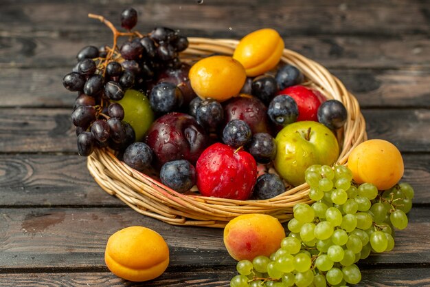 Widok z przodu kosza z owocami łagodnymi i kwaśnymi, takimi jak winogrona, morele, śliwki, na brązowym rustykalnym biurku
