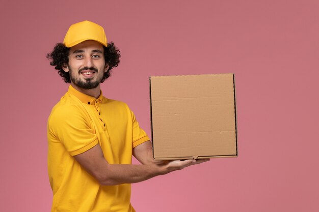 Widok z przodu kurier w żółtym mundurze, trzymający pudełko z dostawą jedzenia na jasnoróżowej ścianie