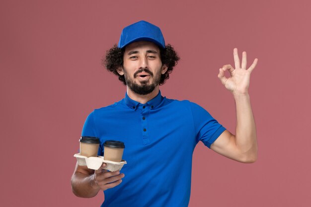 Widok z przodu kuriera w niebieskim mundurze i czapce z filiżankami kawy dostawy na rękach na różowej ścianie