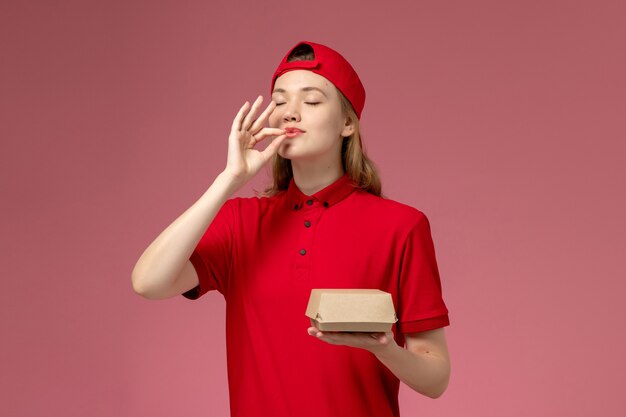 Widok z przodu kurierka w czerwonym mundurze i pelerynie trzymająca małą paczkę z dostawą żywności na różowej ścianie, firma kurierska jednolita dziewczyna praca