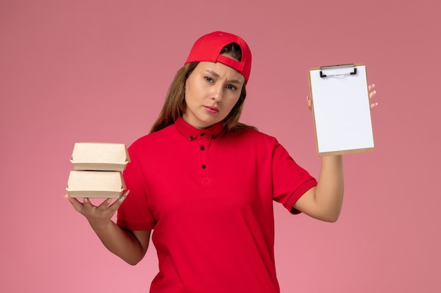 Widok z przodu kurierka w czerwonym mundurze i pelerynie trzymająca mały notatnik z dostawą żywności na jasnoróżowej ścianie, jednolity pracownik biura dostawy pracy