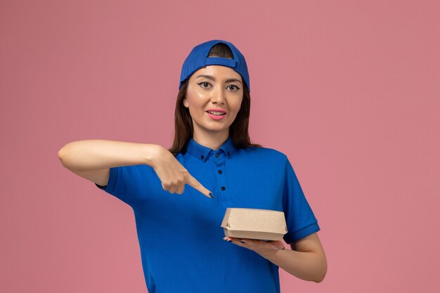 Widok z przodu kurierka w niebieskiej pelerynie munduru trzymająca małą paczkę na różowej ścianie, firma świadcząca usługi dla pracowników