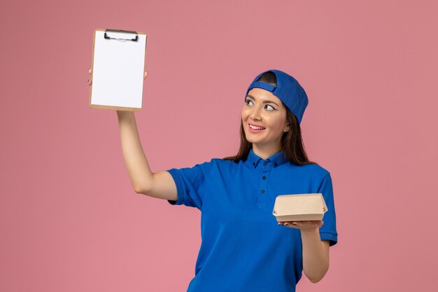Widok z przodu kurierka w niebieskiej pelerynie munduru trzymająca pusty mały pakiet dostawy z notatnikiem na różowej ścianie, dostawa przez firmę usługową dla pracowników