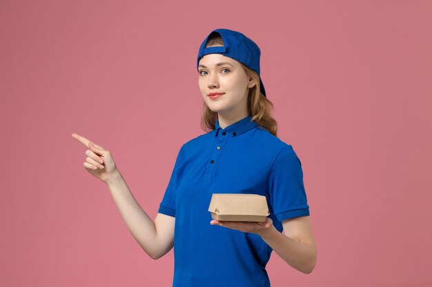 Widok z przodu kurierka w niebieskim mundurze i pelerynie trzymająca małą paczkę z dostawą żywności na różowej ścianie, mundurowa firma usługowa pracownik dziewczyna praca