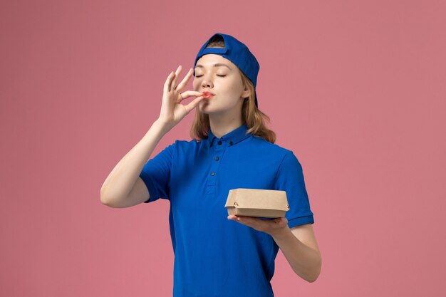 Widok z przodu kurierka w niebieskim mundurze i pelerynie trzymająca małą paczkę z dostawą żywności na różowej ścianie, praca w mundurze dostawy