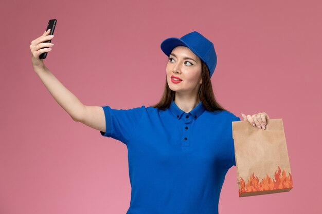 Widok z przodu kurierka w niebieskim mundurze i pelerynie trzymającej telefon i pakiet żywności, robiąc zdjęcie na różowej ścianie