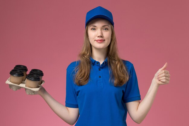 Widok z przodu kurierka w niebieskim mundurze, trzymająca brązowe filiżanki z kawą dostawy z lekkim uśmiechem na różowym biurku jednolity pracownik firmy