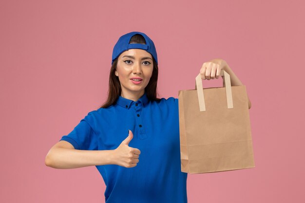 Widok z przodu kurierka w niebieskim mundurze, trzymająca pakiet dostawy papieru na jasnoróżowej ścianie, pracownik serwisu dostarczający kobietę