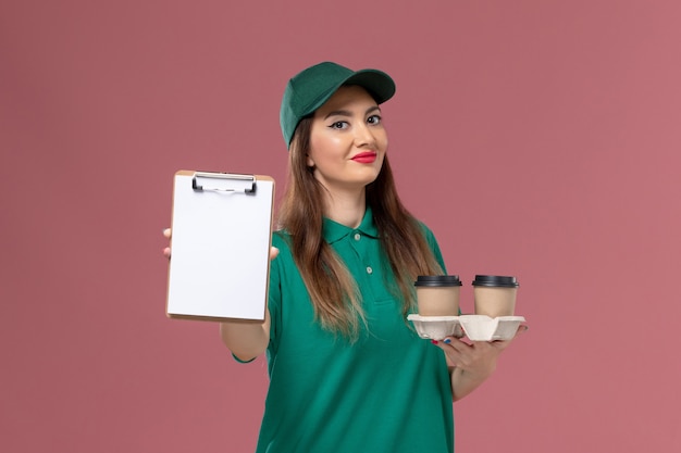 Widok z przodu kurierka w zielonym mundurze i pelerynie trzymająca dostawy filiżanek do kawy i notatnik na różowej ścianie praca pani służba jednolita dostawa