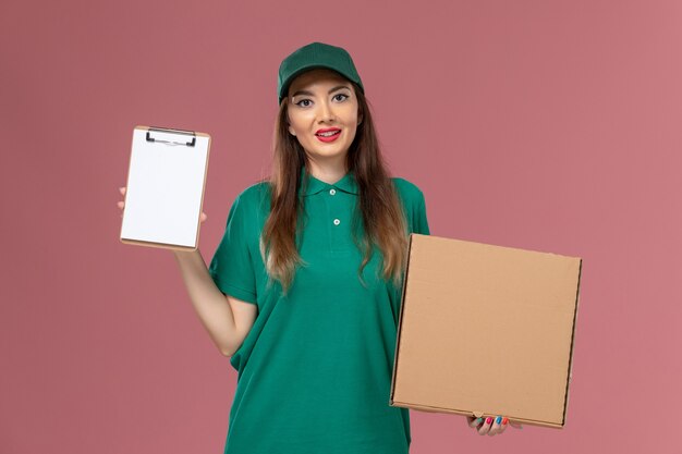 Widok z przodu kurierka w zielonym mundurze, trzymająca pudełko z dostawą żywności i notatnik na różowym biurku, jednolita usługa dostawy
