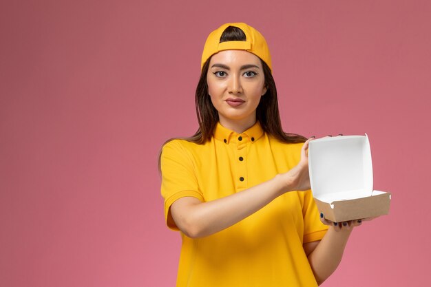 Widok z przodu kurierka w żółtym mundurze i pelerynie trzymająca małą paczkę z dostawą żywności i otwierająca ją na różowej ścianie pracownik firmy świadczącej usługi w mundurze