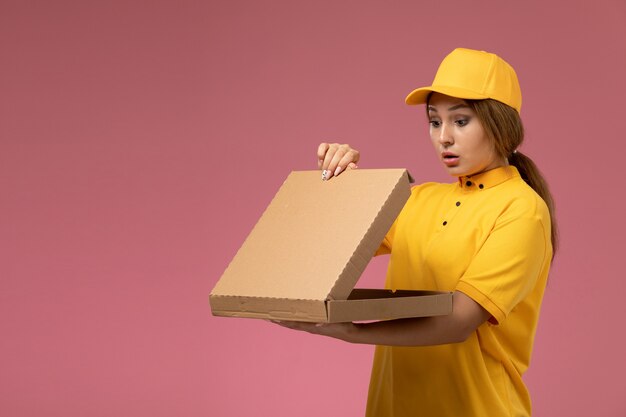 Widok z przodu kurierka w żółtym mundurze żółtej peleryny trzymającej i otwierającej pudełko z jedzeniem na różowym biurku jednolita dostawa kobiecego koloru