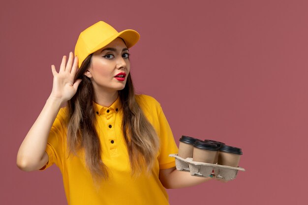 Widok z przodu kurierki w żółtym mundurze i czapce trzymającej brązowe filiżanki z kawą i próbującej usłyszeć na różowej ścianie