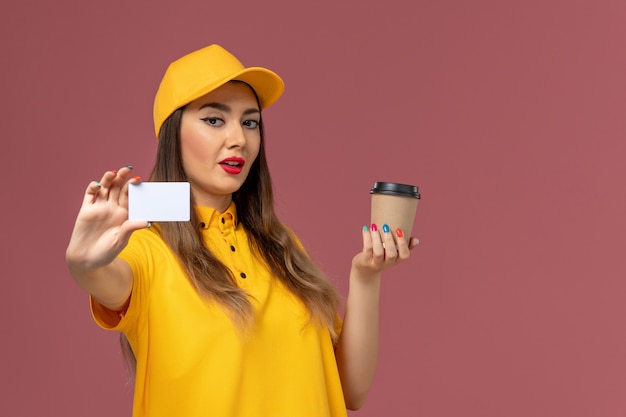 Widok z przodu kurierki w żółtym mundurze i czapce trzymającej dostawę filiżanki kawy i białą kartkę na różowej ścianie