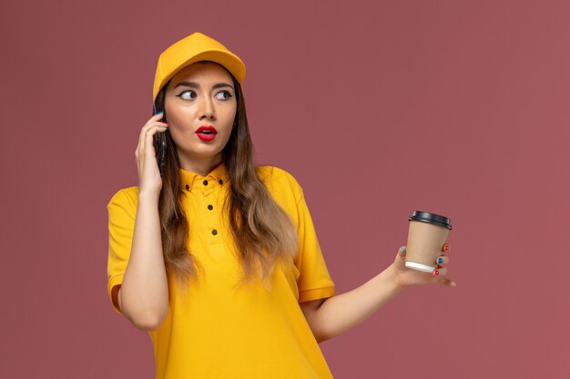 Widok z przodu kurierki w żółtym mundurze i czapce trzymającej filiżankę kawy dostawy i rozmawiającej przez telefon na różowej ścianie