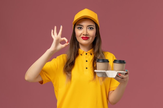 Widok z przodu kurierki w żółtym mundurze i czapce trzymającej filiżanki kawy dostawy na różowej ścianie