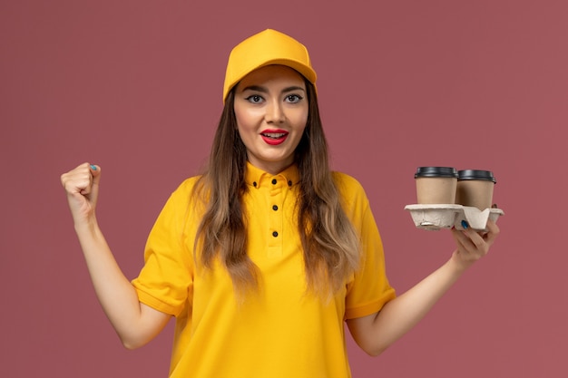 Bezpłatne zdjęcie widok z przodu kurierki w żółtym mundurze i czapce trzymającej filiżanki z kawą dostawy radującą się na różowej ścianie
