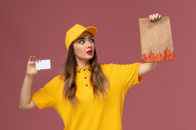 Widok z przodu kurierki w żółtym mundurze i czapce trzymającej opakowanie żywności i plastikową kartę na różowej ścianie