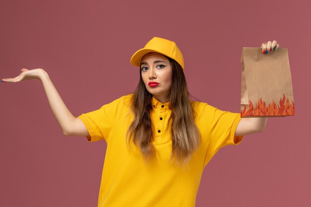 Widok z przodu kurierki w żółtym mundurze i czapce trzymającej paczkę z jedzeniem na jasnoróżowej ścianie