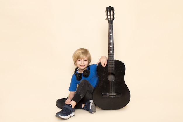 Bezpłatne zdjęcie widok z przodu ładny mały chłopiec w niebieskiej koszulce z czarnymi słuchawkami z czarną gitarą