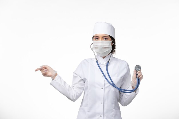 Widok z przodu lekarka w białym sterylnym kombinezonie medycznym i noszącej maskę z powodu COVID - używanie stetoskopu na białej ścianie choroba wirus - choroba pandemiczna
