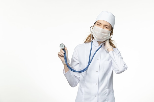 Widok z przodu lekarka w kombinezonie medycznym nosząca maskę z powodu koronawirusa trzymającego stetoskop na jasnej białej ścianie wirusa pandemii wirusa - choroba