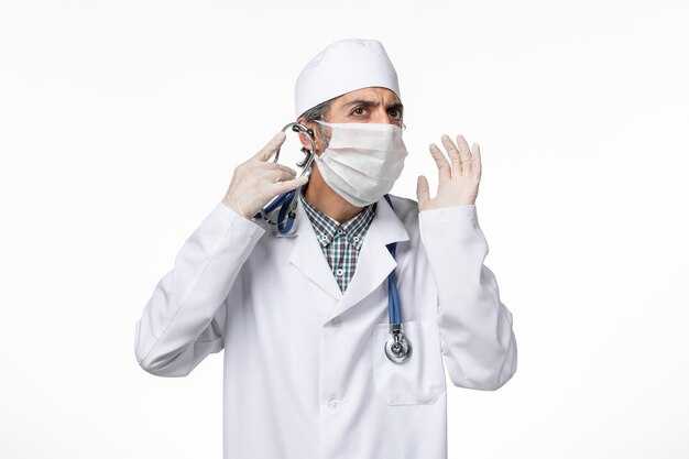 Widok z przodu lekarz mężczyzna w białym kombinezonie medycznym w masce z powodu koronawirusa na jasnobiałej powierzchni