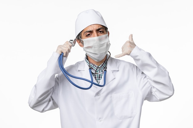 Bezpłatne zdjęcie widok z przodu lekarz mężczyzna w białym kombinezonie medycznym w masce z powodu koronawirusa trzymającego stetoskop na białej powierzchni