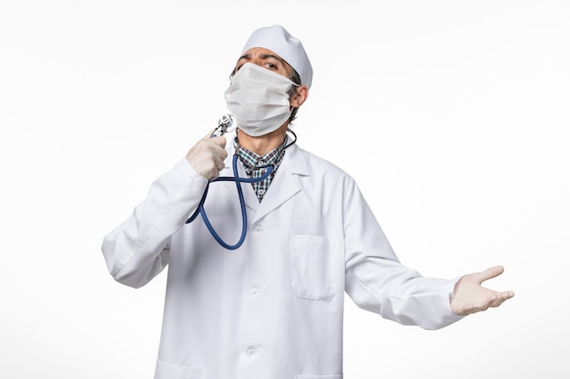 Bezpłatne zdjęcie widok z przodu lekarz mężczyzna w białym kombinezonie medycznym w masce z powodu koronawirusa trzymającego stetoskop na białym biurku
