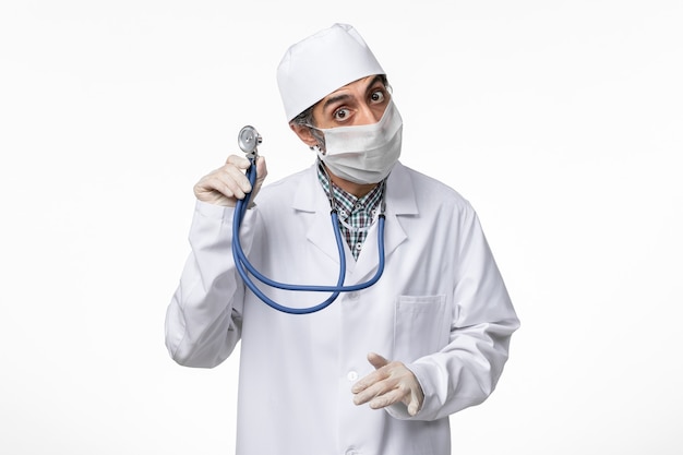 Widok z przodu lekarz mężczyzna w białym kombinezonie medycznym w masce z powodu koronawirusa trzymającego stetoskop na jasnobiałej powierzchni