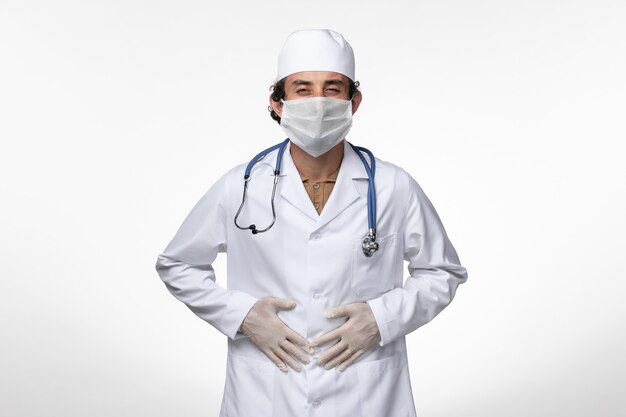 Widok z przodu lekarza płci męskiej w kombinezonie medycznym i noszącym maskę jako ochrona przed chorobą zakaźną - cierpiący na ból brzucha na białej ścianie choroba wirus - pandemia