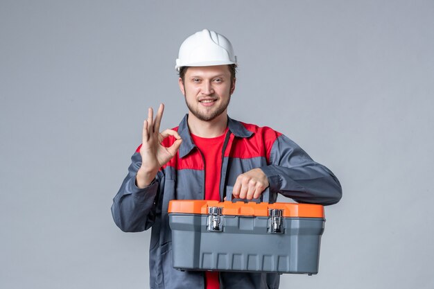 widok z przodu męski budowniczy w mundurze i kasku trzymającym walizkę narzędziową na szarym tle