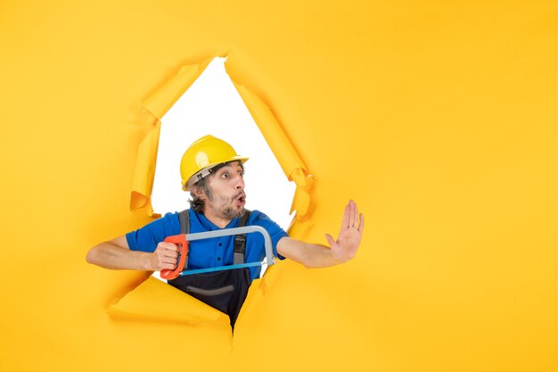 Bezpłatne zdjęcie widok z przodu męski budowniczy w mundurze trzymającym piłę łukową na żółtym tle