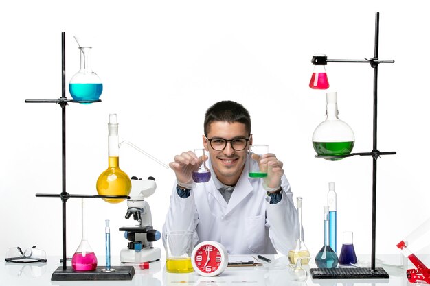 Widok z przodu męski chemik w białym kombinezonie medycznym trzymający kolby z roztworami na jasnobiałym tle wirus naukowy COVID - laboratorium pandemiczne