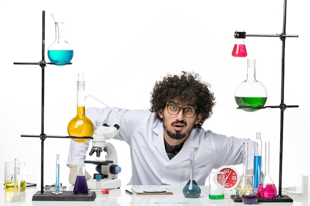 Bezpłatne zdjęcie widok z przodu męski chemik w garniturze medycznym, siedzący przed stołem z roztworami na białej przestrzeni