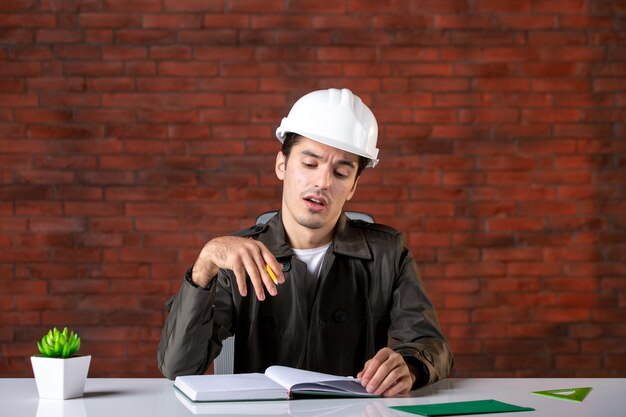 Widok z przodu męski inżynier siedzący za swoim miejscem pracy w białym kasku dokument biznes własność korporacyjna program pracy konstruktor plan wykonawcy