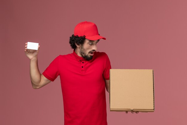 Widok z przodu męski kurier w czerwonej koszuli i pelerynie trzymający pudełko z jedzeniem dostawy i kartę na różowej ścianie