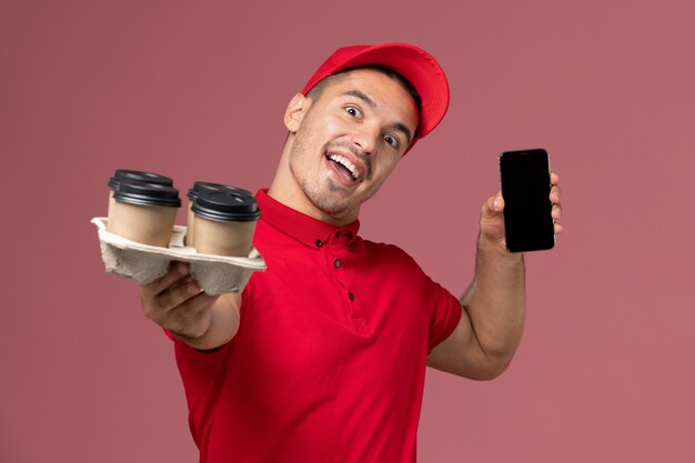 Widok z przodu męski kurier w czerwonym mundurze i pelerynie, trzymając filiżanki kawy dostawy z telefonem na różowej ścianie