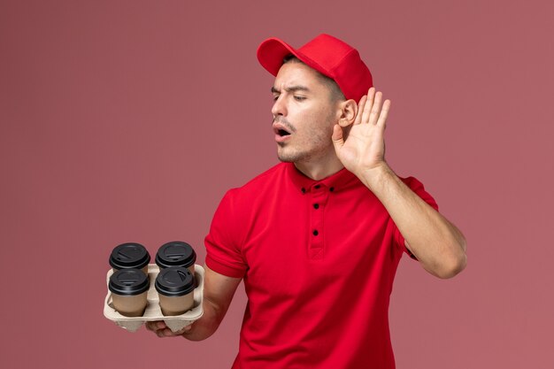Widok z przodu męski kurier w czerwonym mundurze trzymający brązowe filiżanki kawy dostawy próbujący usłyszeć na jasnoróżowym pracowniku ściennym