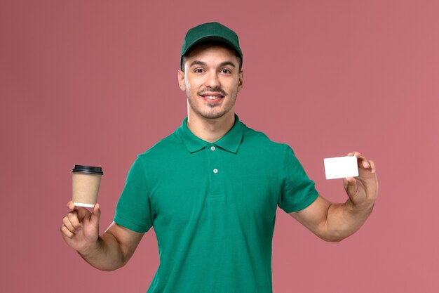 Widok z przodu męski kurier w zielonym mundurze, trzymając filiżankę kawy dostawy z białą kartą na jasnoróżowym tle